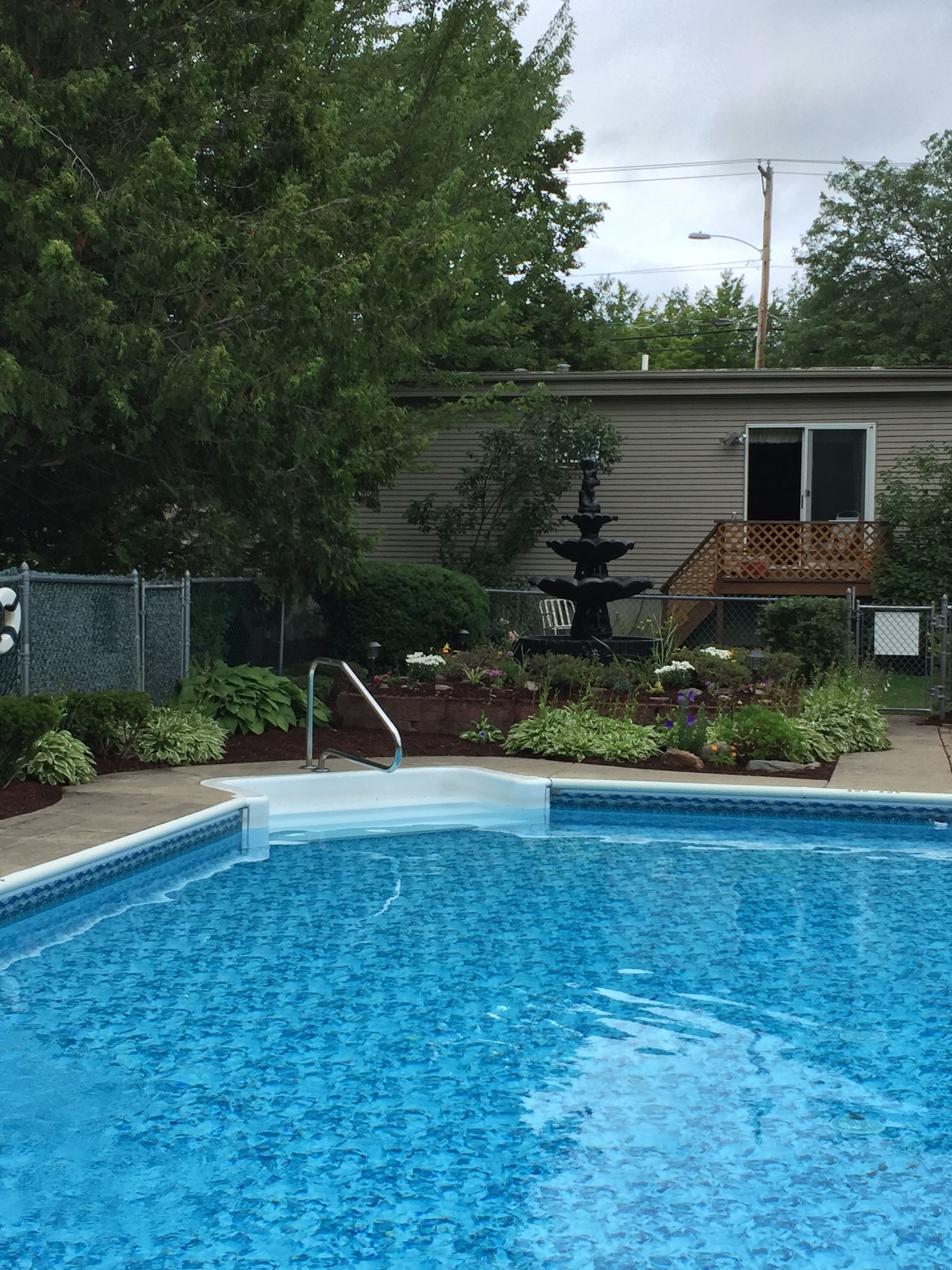 Une piscine avec un escalier, entourée d’une clôture, d’arbres et d’une maison dotée d’une terrasse.