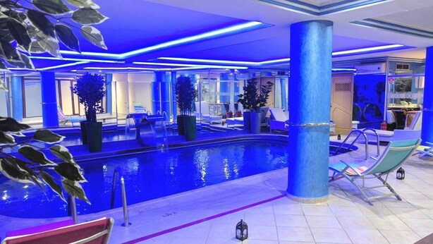 An indoor swimming pool area with blue lighting, lounge chairs, and potted plants. - Hotel Rabat (Rabat)