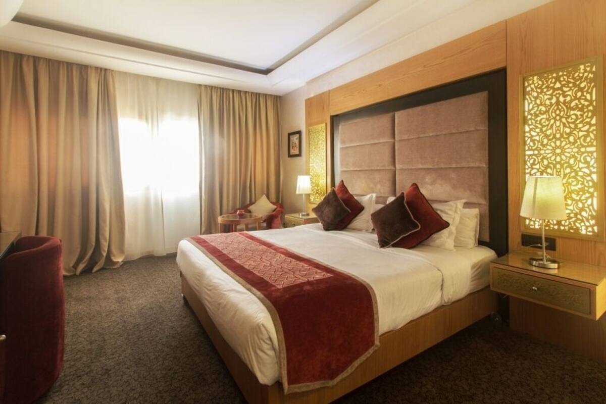 A hotel room with a large bed, a wooden headboard, a red throw blanket, a nightstand with a lamp, and a red chair.