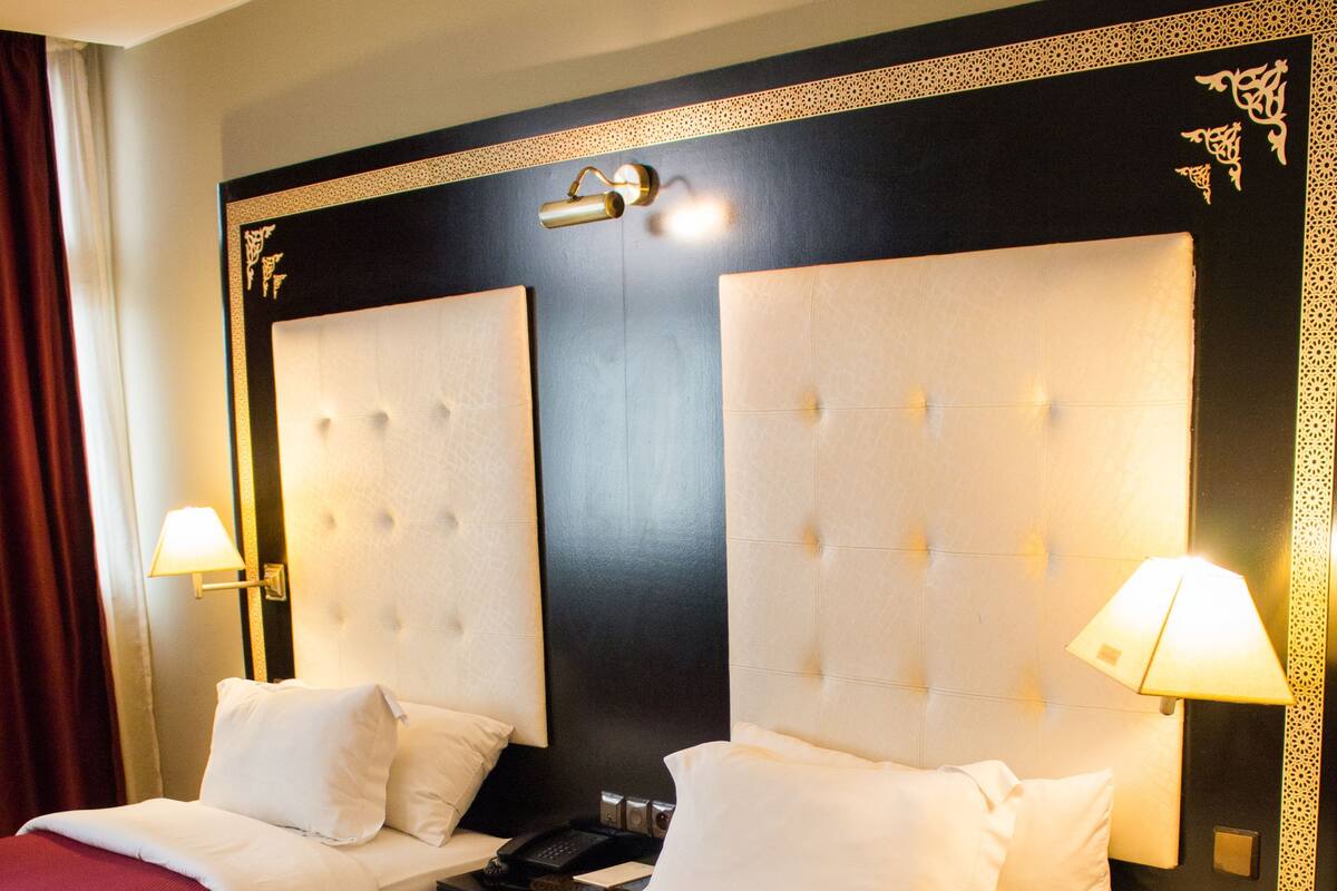 A hotel room with two beds, each with a white headboard and red bedding. There are bedside tables with lamps, a nightstand, and a small table with a lamp.