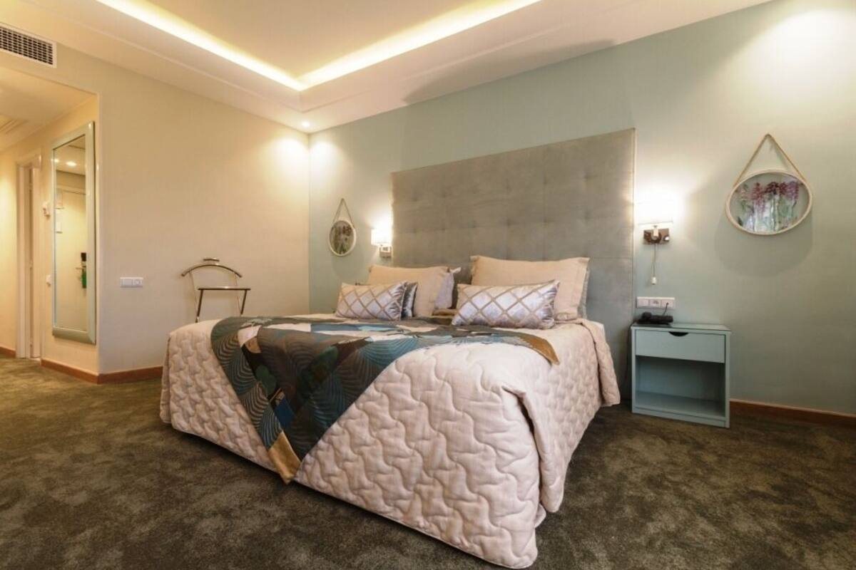 A bedroom with a large bed, a bedside table, a wall-mounted lamp, and a decorative mirror.