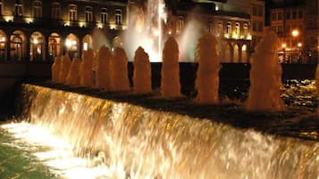 Vista nocturna de una fuente con chorros de agua, rodeada de edificios con ventanas iluminadas.