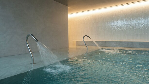 Een modern binnenzwembad met twee waterstralen, een betegelde wand en een warme lichtbalk.