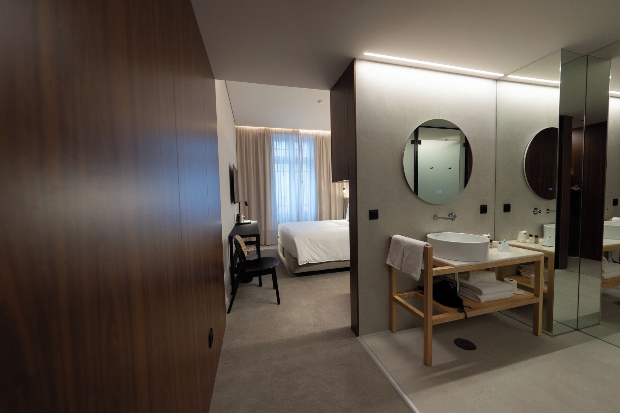 Une chambre d’hôtel moderne équipée d’un lit, d’un bureau, d’un miroir rond et d’une coiffeuse avec un lavabo.