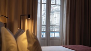 Een hotelkamer met een bed, nachtkastje, lamp en uitzicht op een gebouw door het raam.