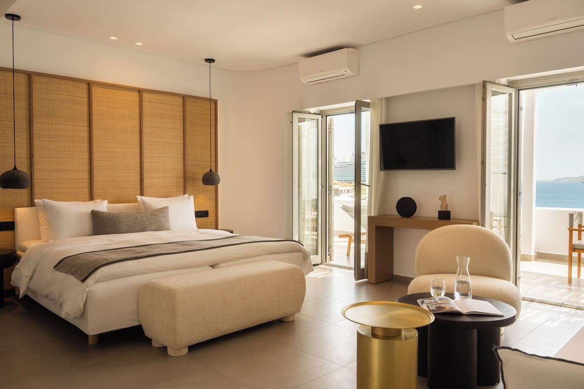 A modern hotel room with a large bed, a sitting area with a chair and a table, a flat-screen TV, and a balcony with a view of the sea.