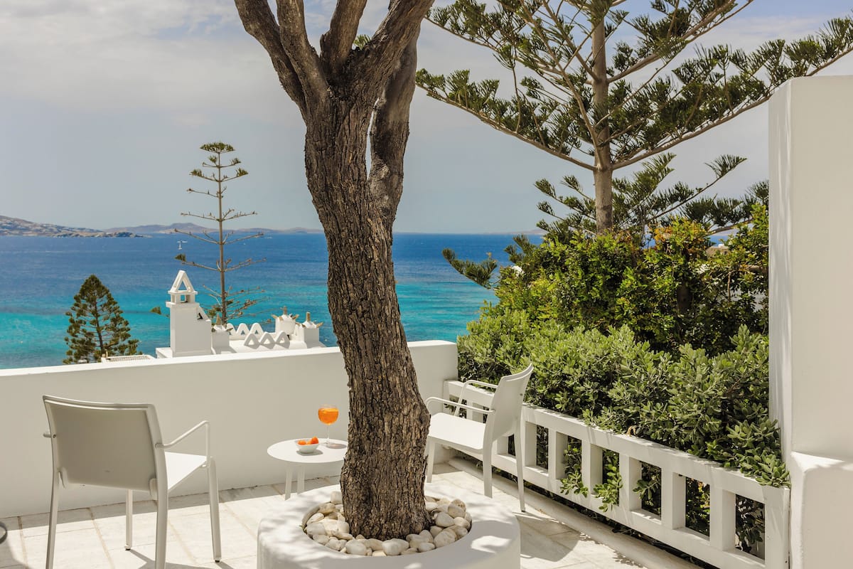 A balcony with a tree, white chairs, a table, and a view of the sea.