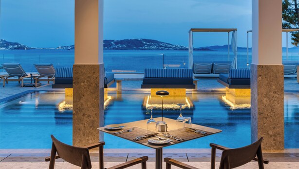A poolside dining area with a table set for two, overlooking a pool with a view of the sea and distant hills. - Grace Mykonos (Mykonos)