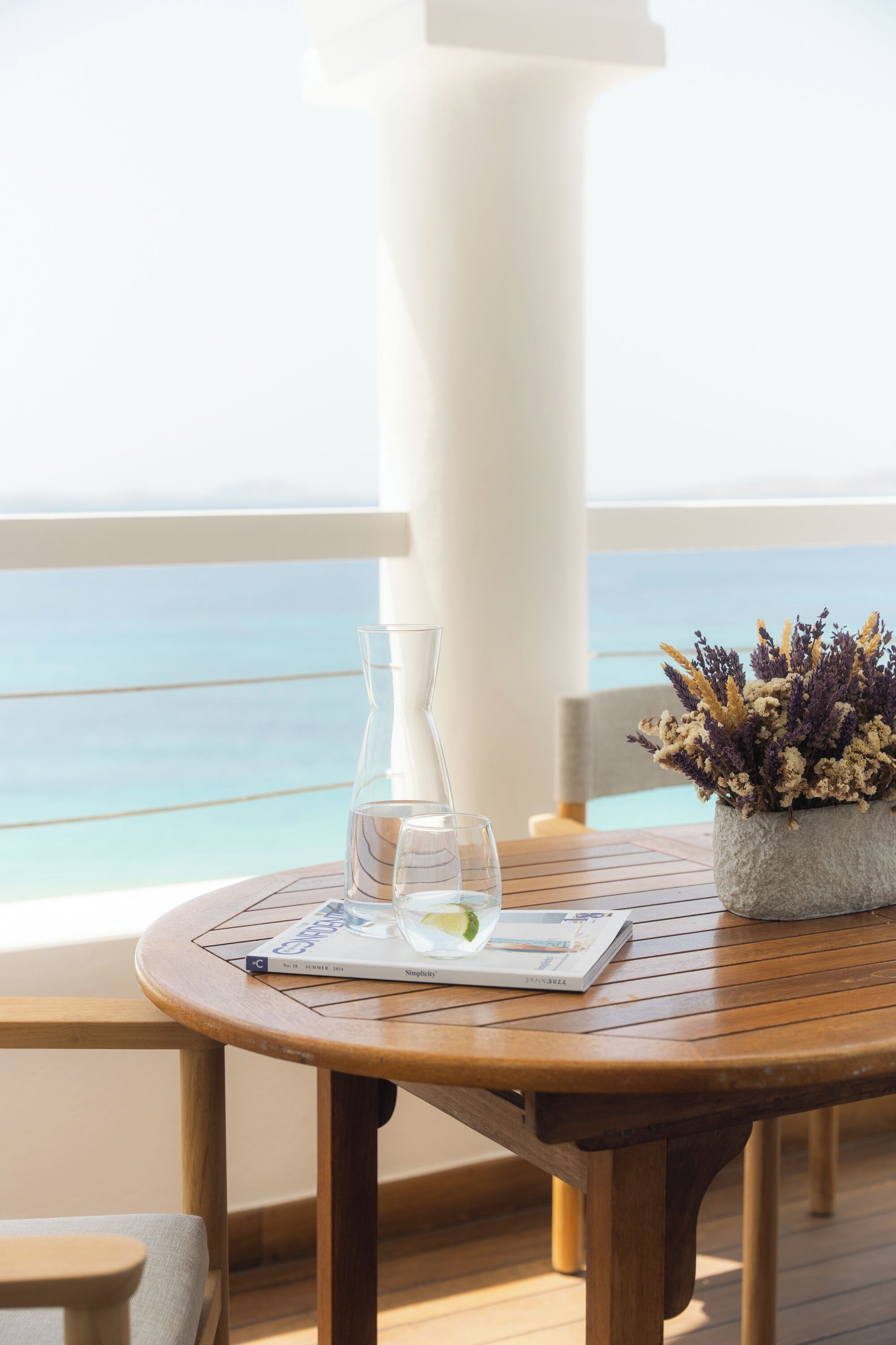 A wooden table with a glass decanter, a glass, and a menu on a balcony overlooking the sea.