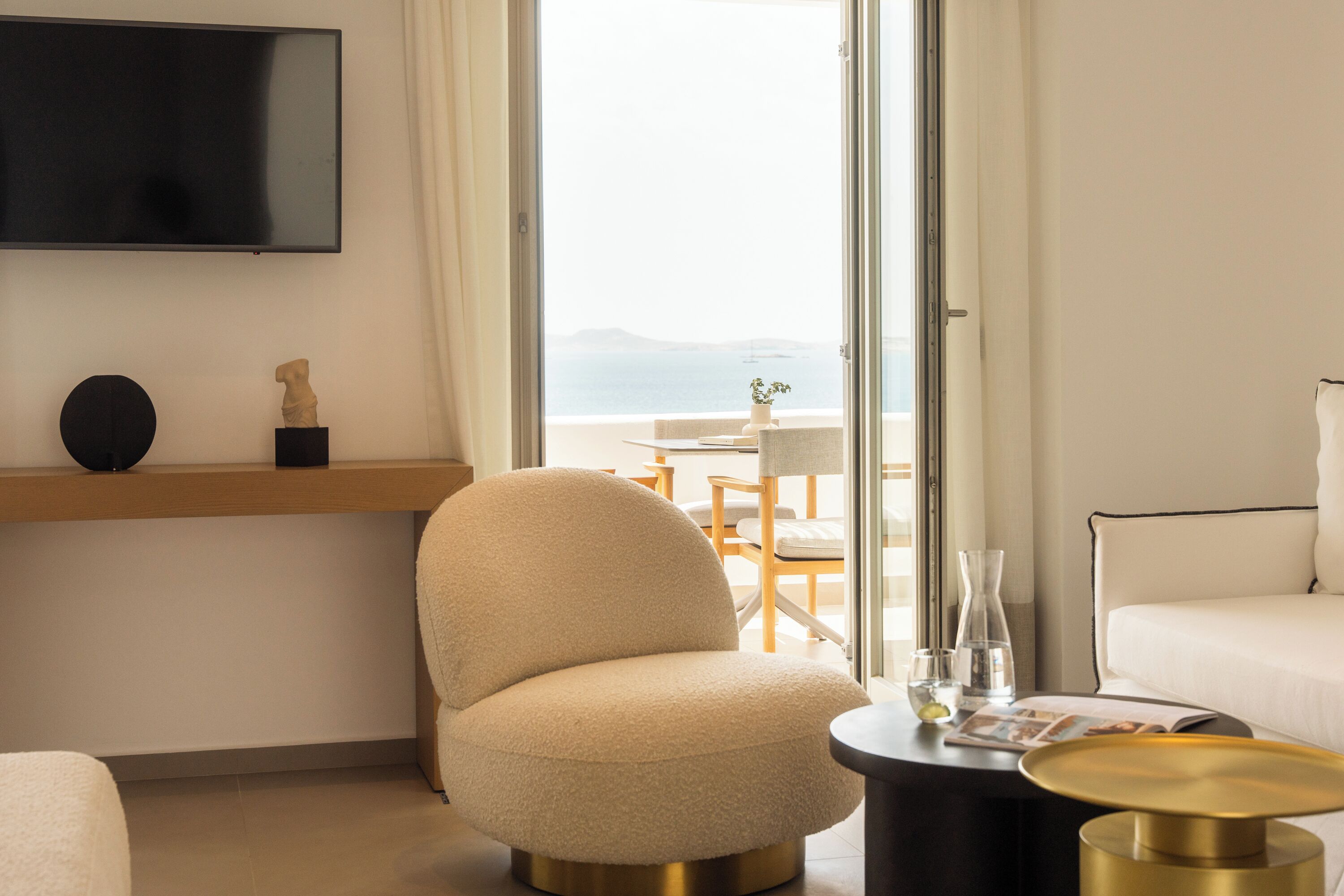 Habitación de hotel moderna con sofá, un sillón redondo, una mesita de centro, un televisor de pantalla plana en la pared y vista al mar a través de un ventanal grande.