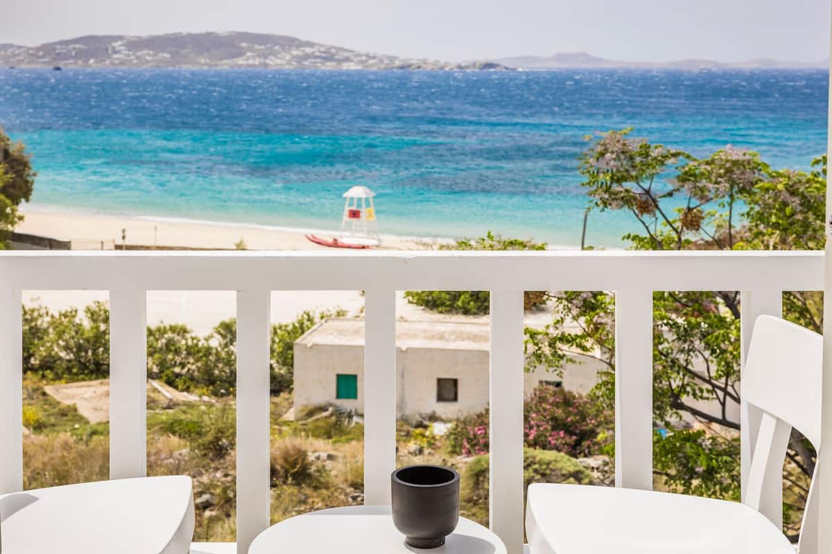 A balcony with white chairs, a table, and a view of a beach with a lighthouse.