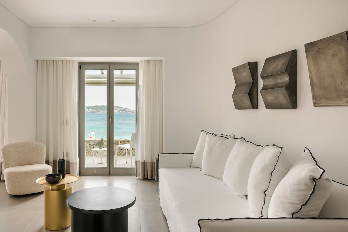 A modern living room with a white sofa, a round black coffee table, and a beige armchair. There are abstract wall art pieces and a view of the sea through a glass door.