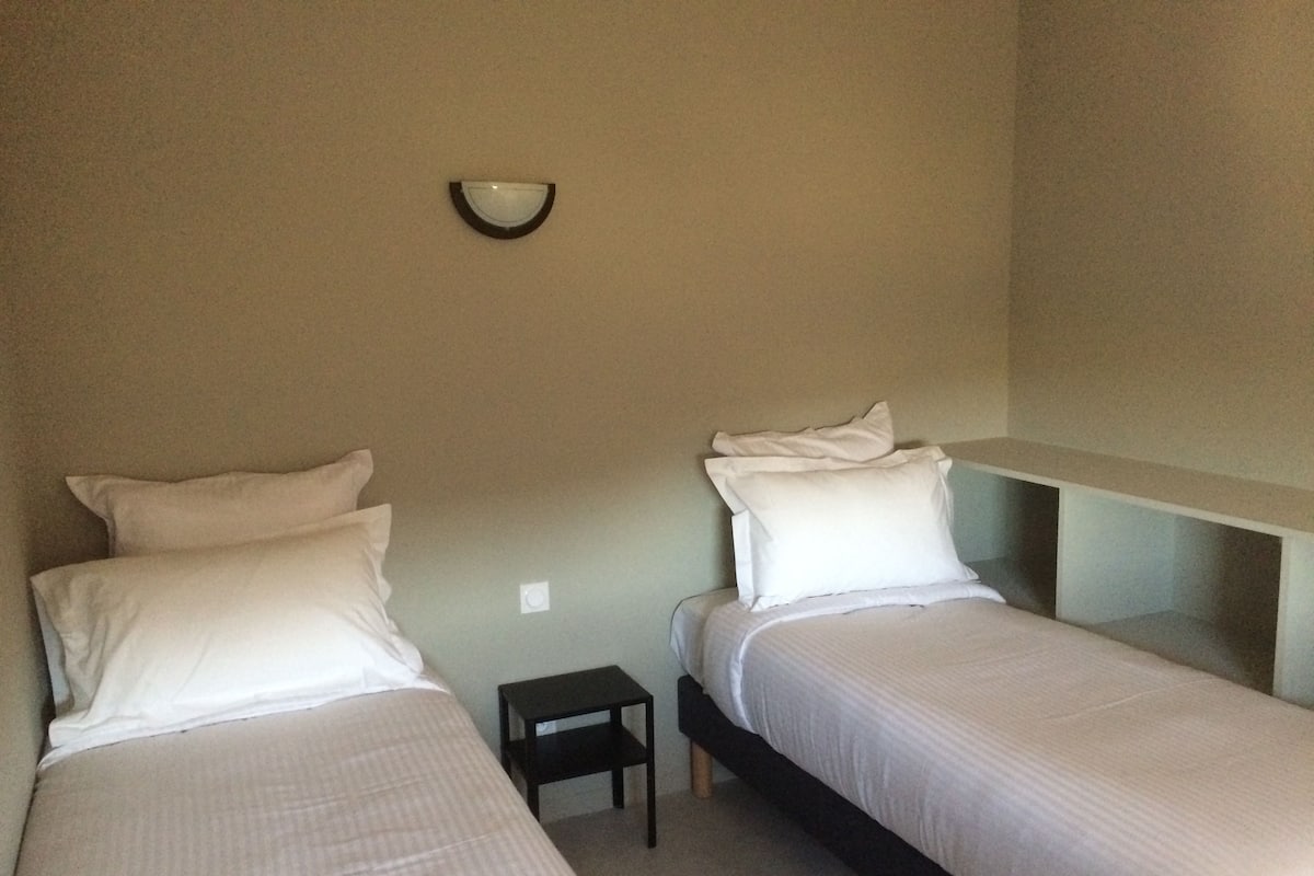 A small, simple room with two single beds, a nightstand, and a wall-mounted light fixture.