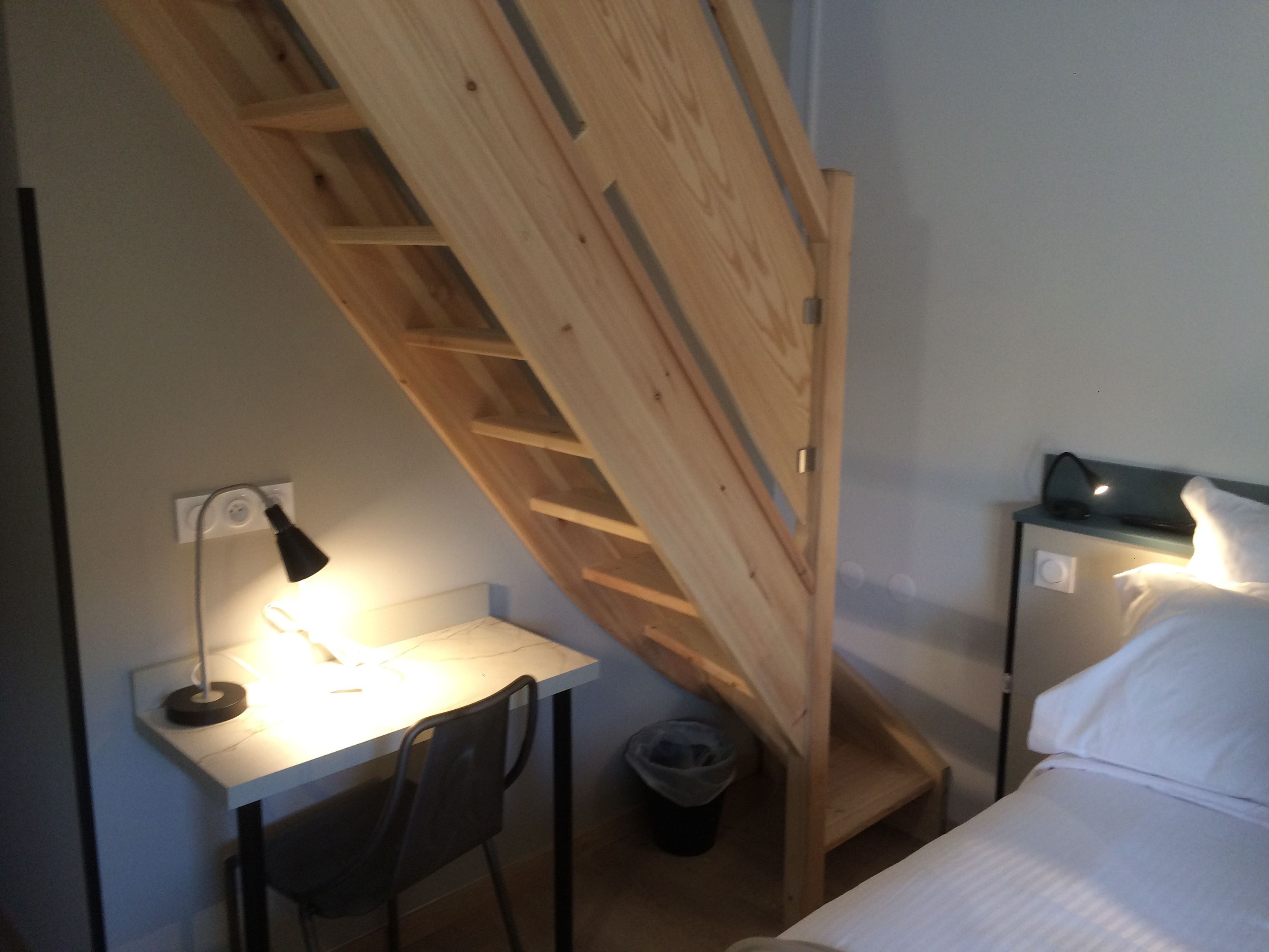 Habitación pequeña con escritorio, silla y cama. Una escalera de madera conduce a una zona superior.