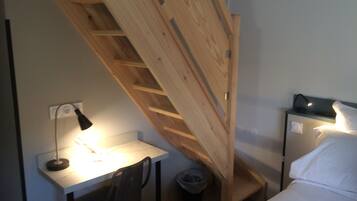 Habitación pequeña con escritorio, silla y cama. Una escalera de madera conduce a una zona superior.