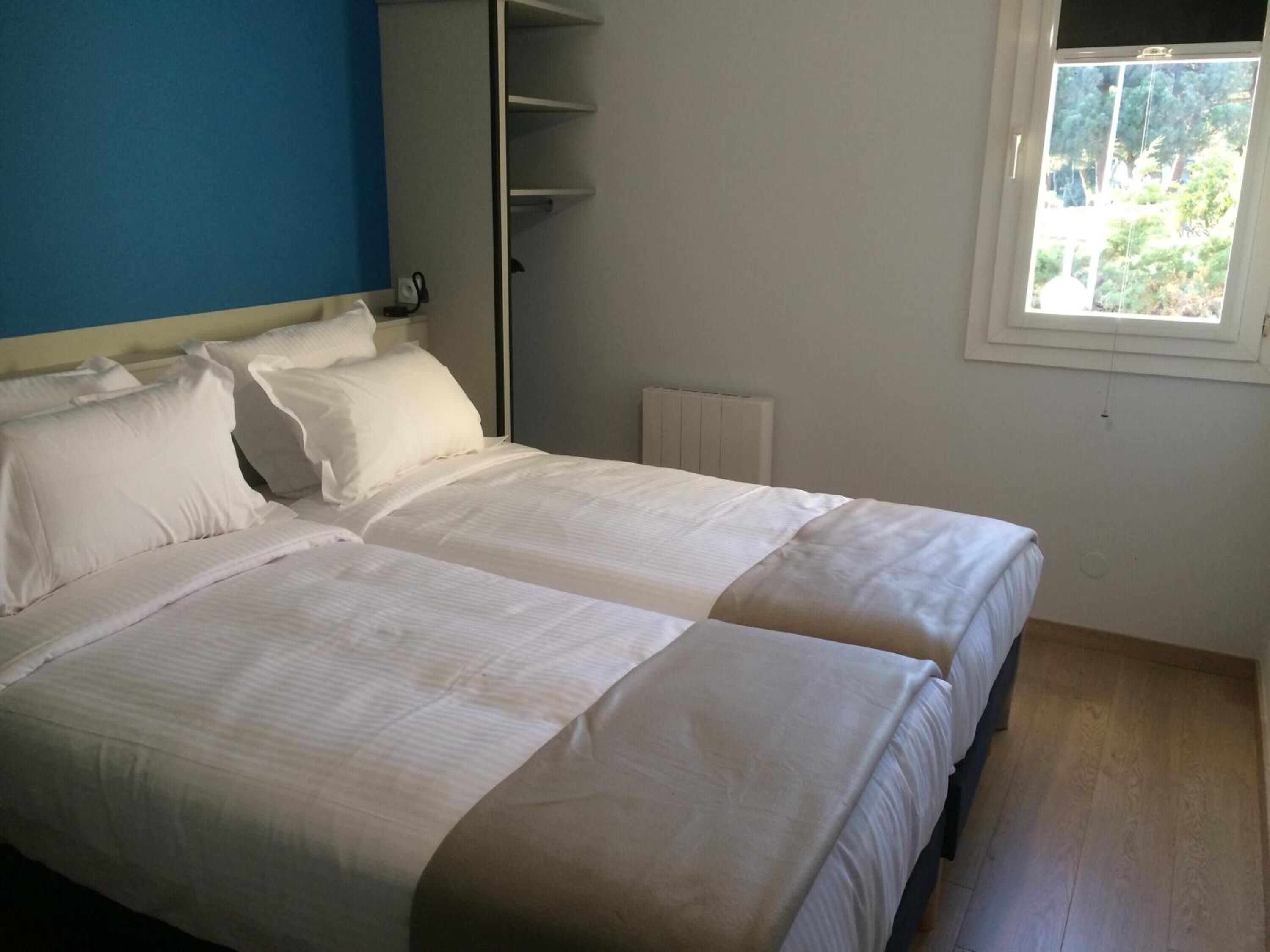 Cama doble con ropa de cama blanca en una habitación con pared azul y ventana con vistas a zonas verdes.