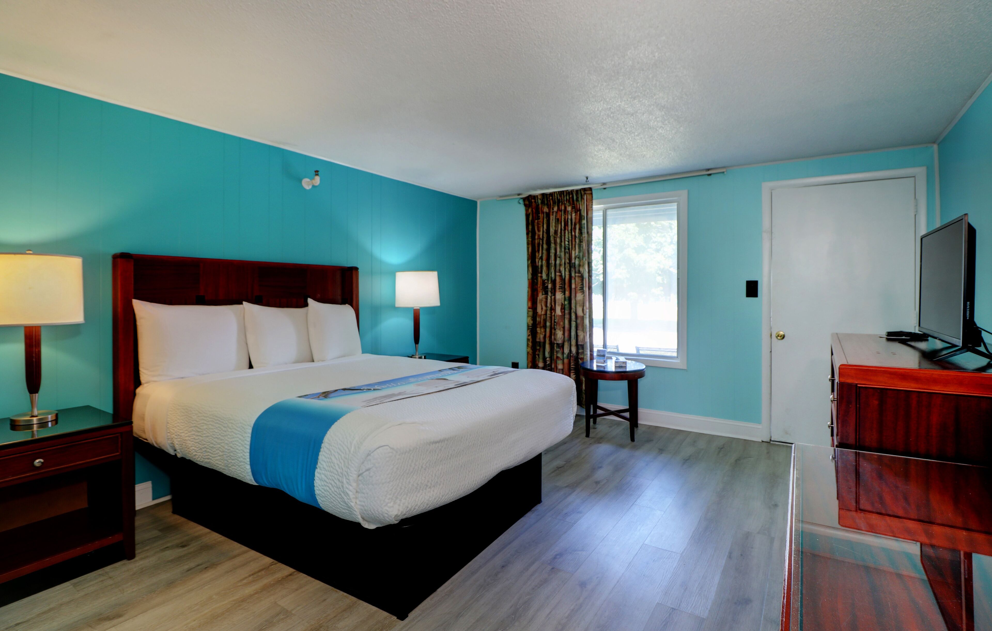 A hotel room with a large bed, two bedside lamps, a desk with a chair, a television, and a window with curtains.