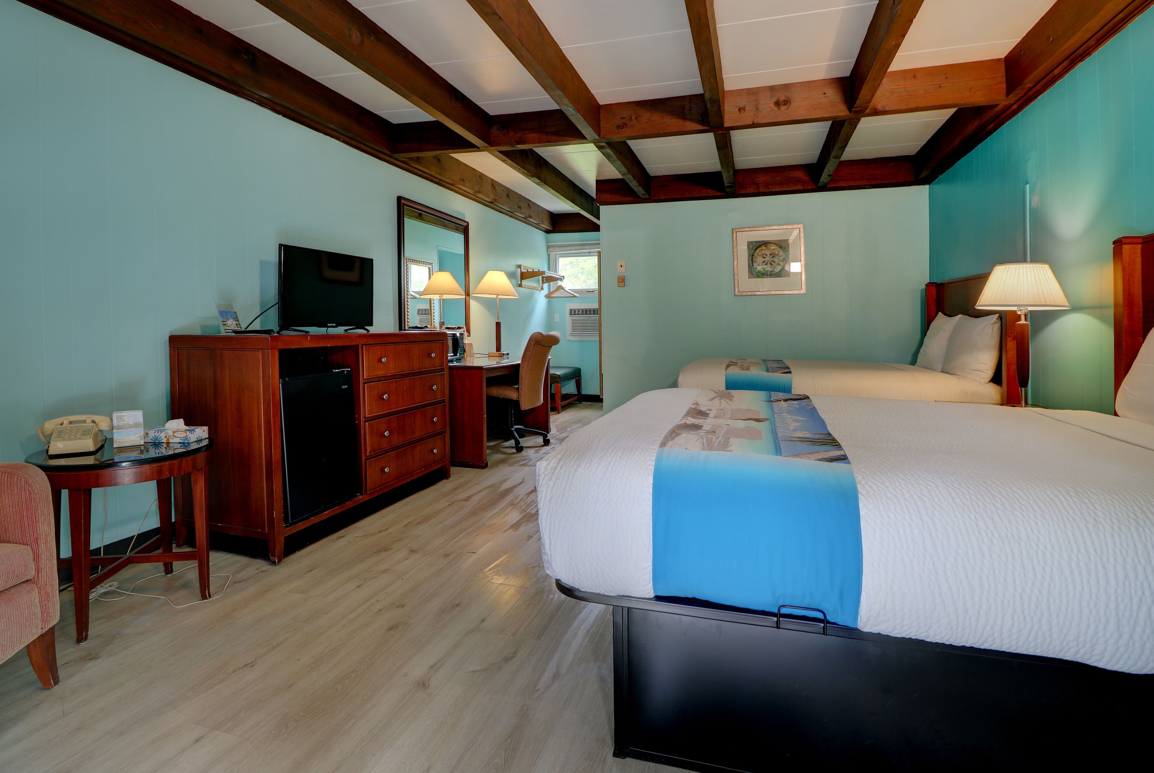 A hotel room with a wooden ceiling, a bed with a blue blanket, a desk with a lamp, a chair, and a television.