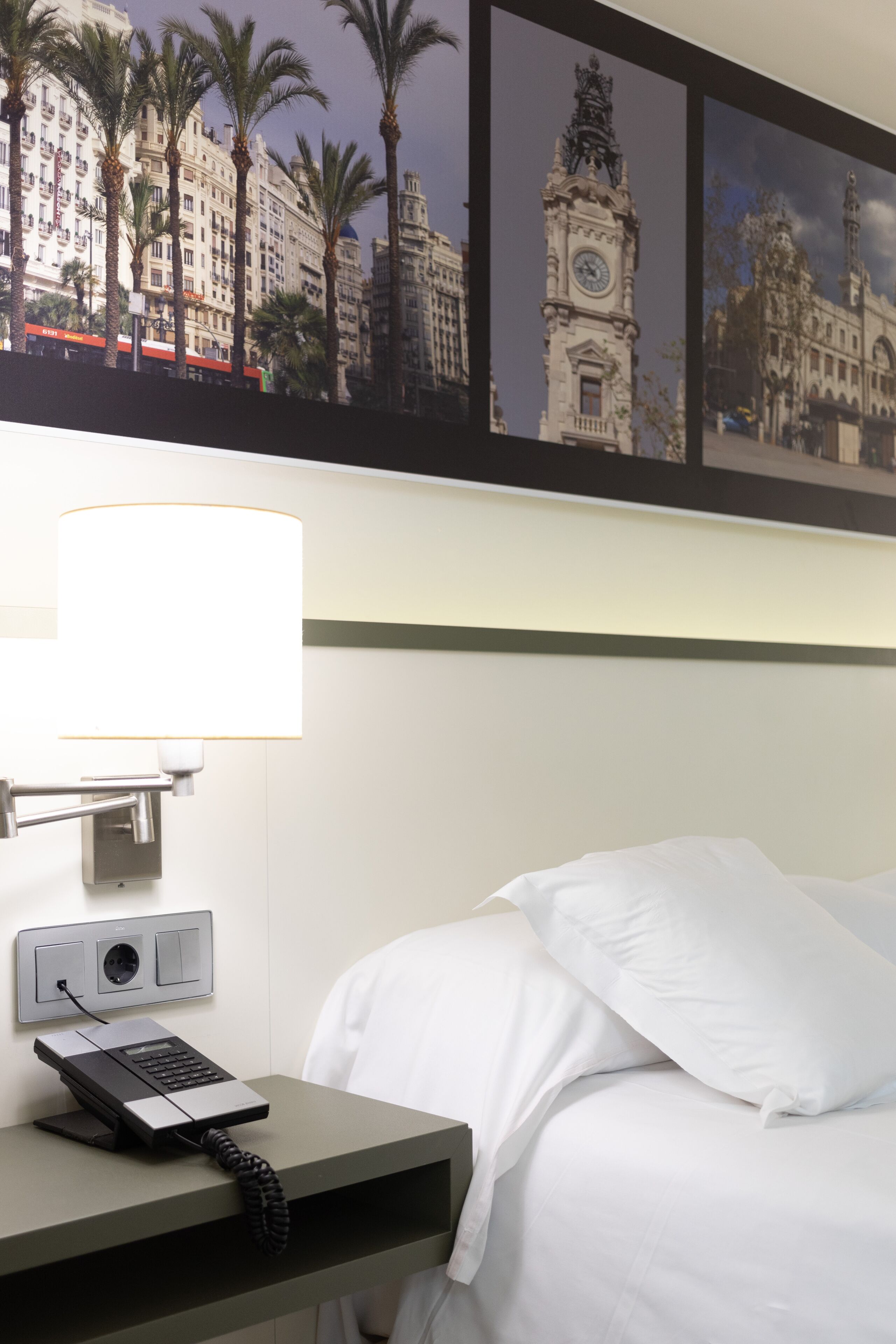 A hotel room with a bed, a bedside table with a lamp, a phone, and a framed cityscape picture on the wall.