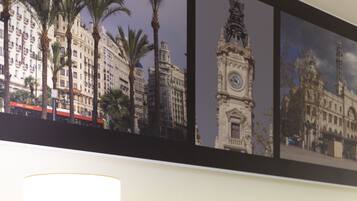 A hotel room with a bed, a bedside table with a lamp, a phone, and a framed cityscape picture on the wall.