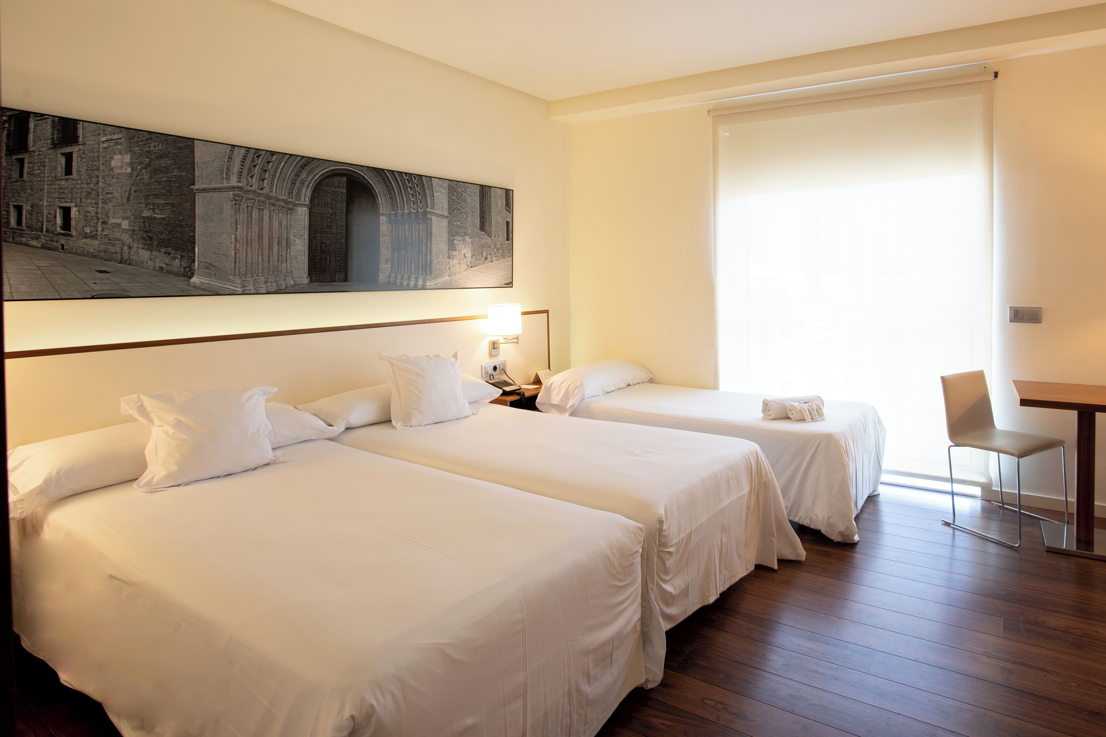 A hotel room with two beds, a desk, a chair, and a large painting on the wall.