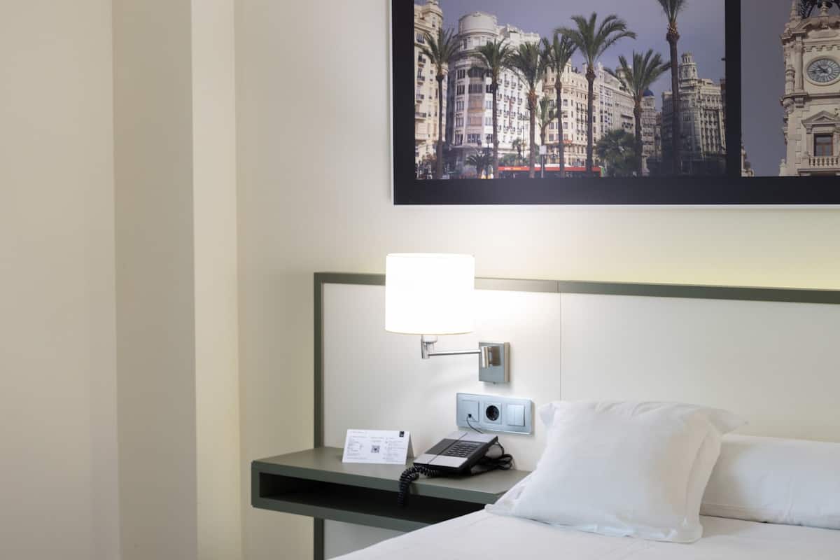 Habitación de hotel con cama, mesita de noche con lámpara, teléfono y televisión de pared mostrando una vista urbana.
