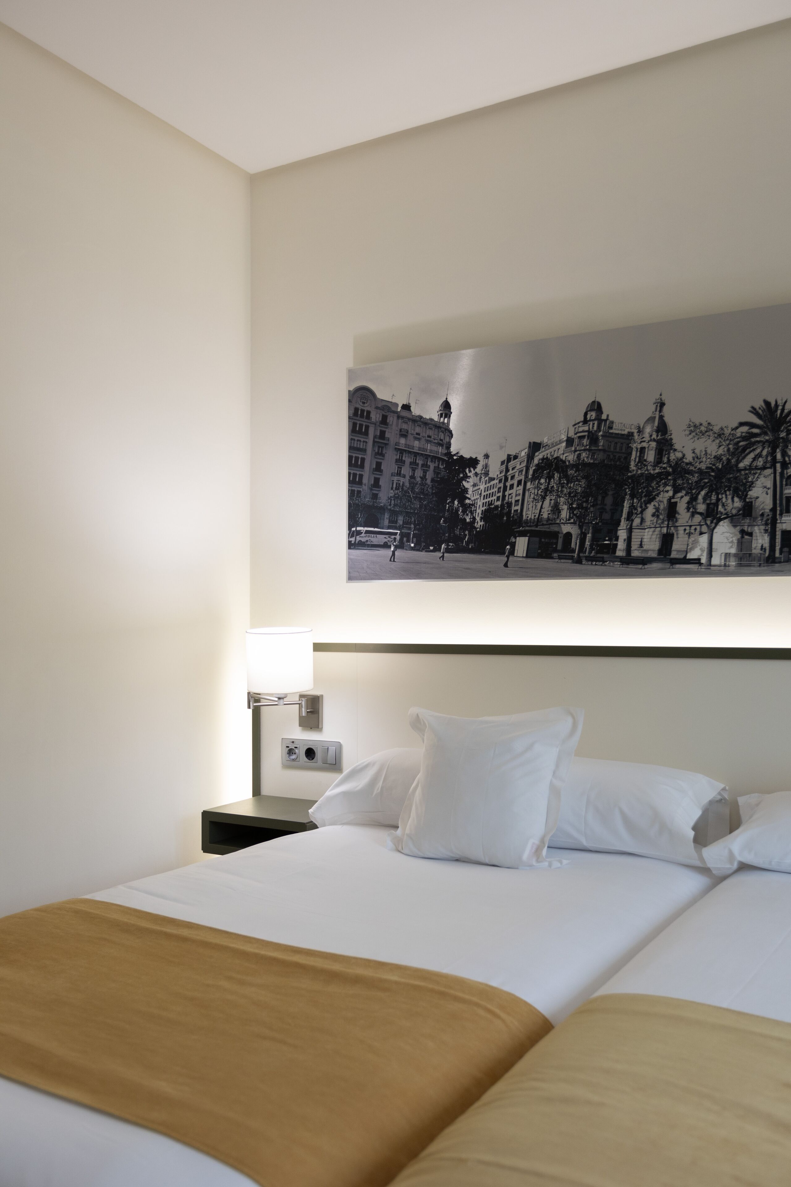 A hotel room with a bed, a nightstand, a lamp, and a wall-mounted picture of a cityscape.