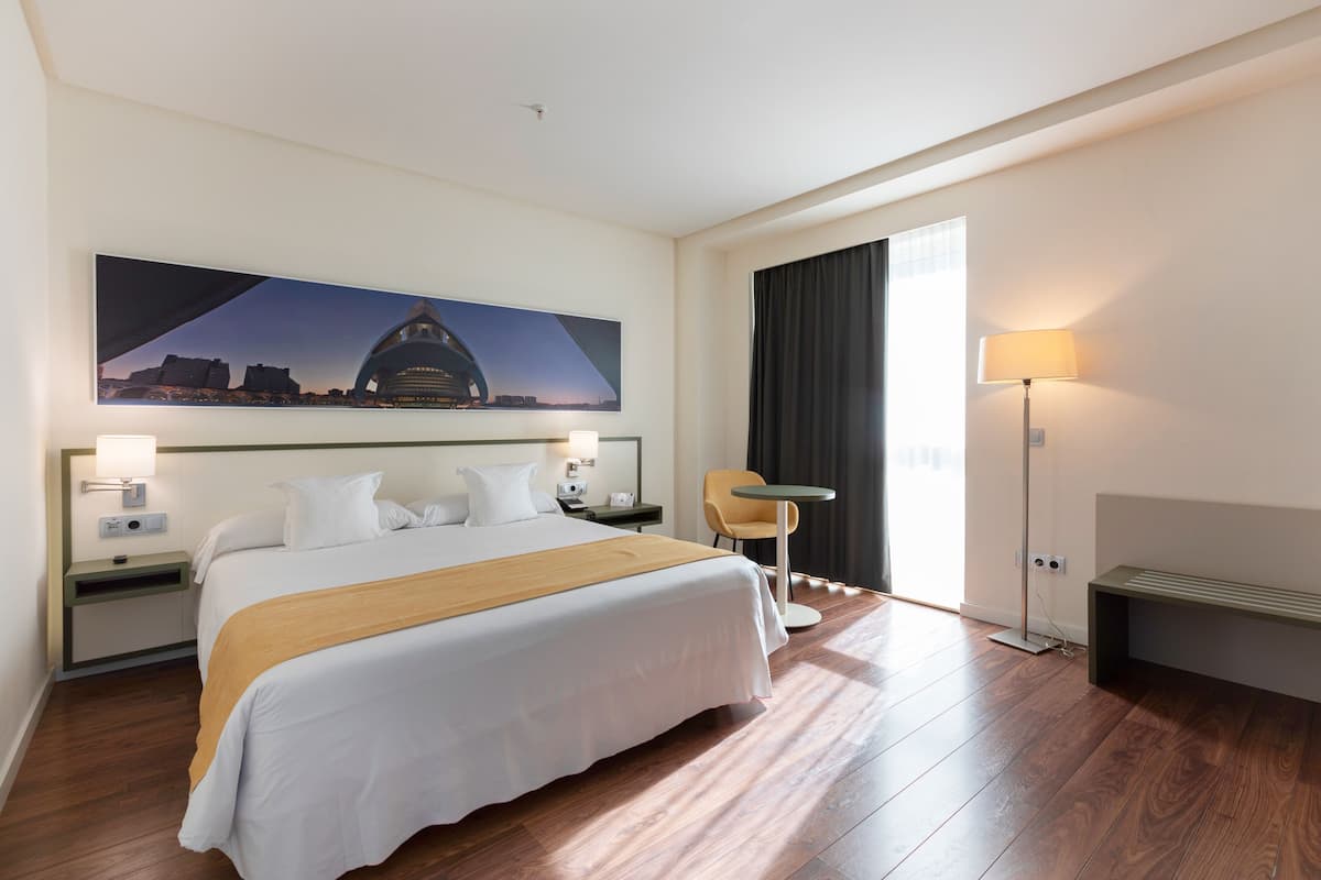 Habitación de hotel con una cama grande, un escritorio, una silla, una lámpara y un ventanal amplio.