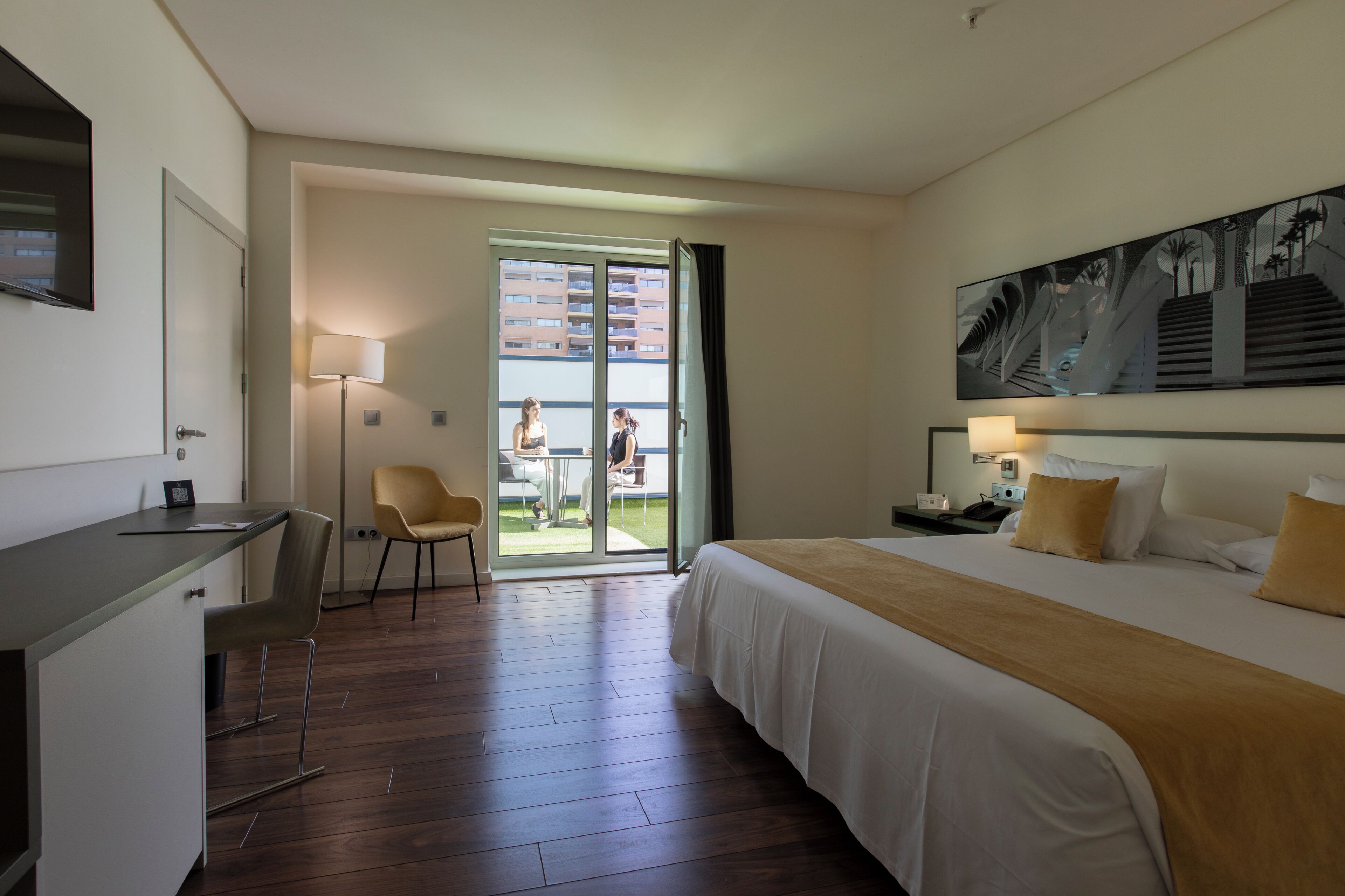 A hotel room with a bed, desk, chair, and a view of the outside balcony.
