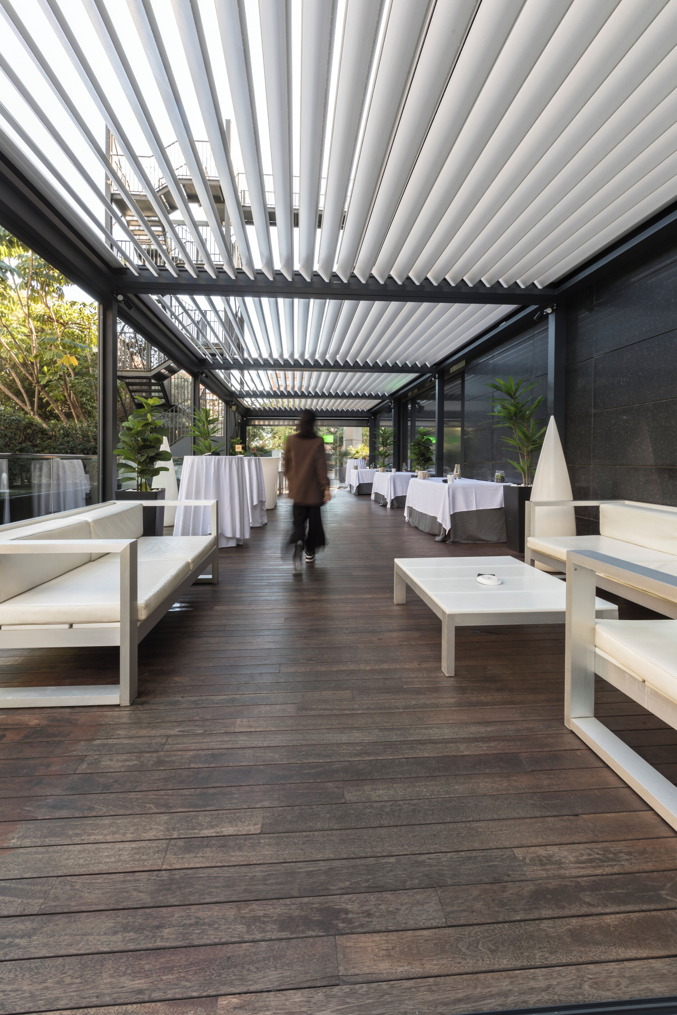 Un espace extérieur abrité avec des tables et des bancs, une personne qui marche, et une vue sur des arbres et des bâtiments en arrière-plan.