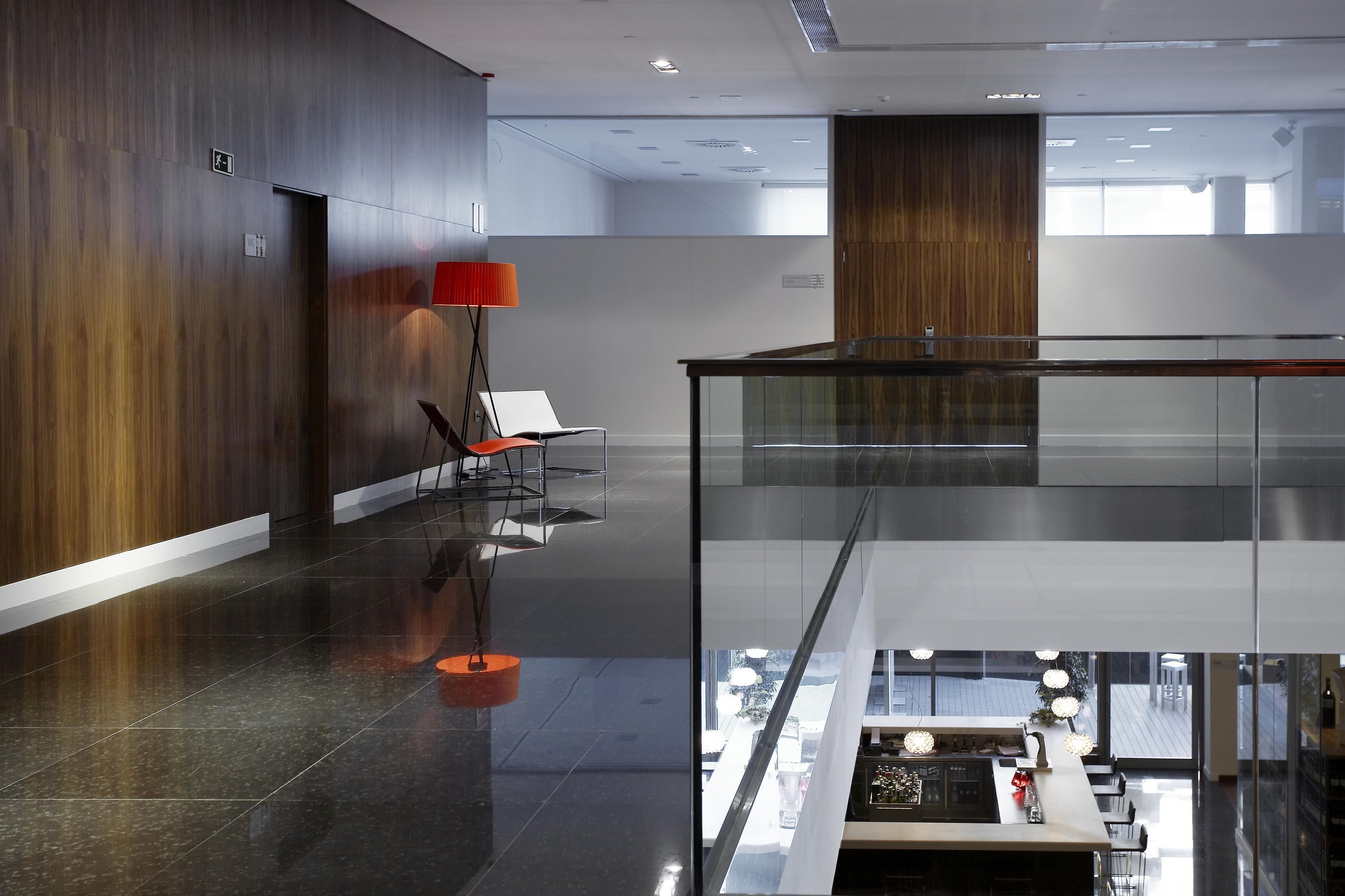 Un lobby moderno de hotel con barandilla de vidrio, una zona de estar con una lámpara roja y vistas al piso inferior.