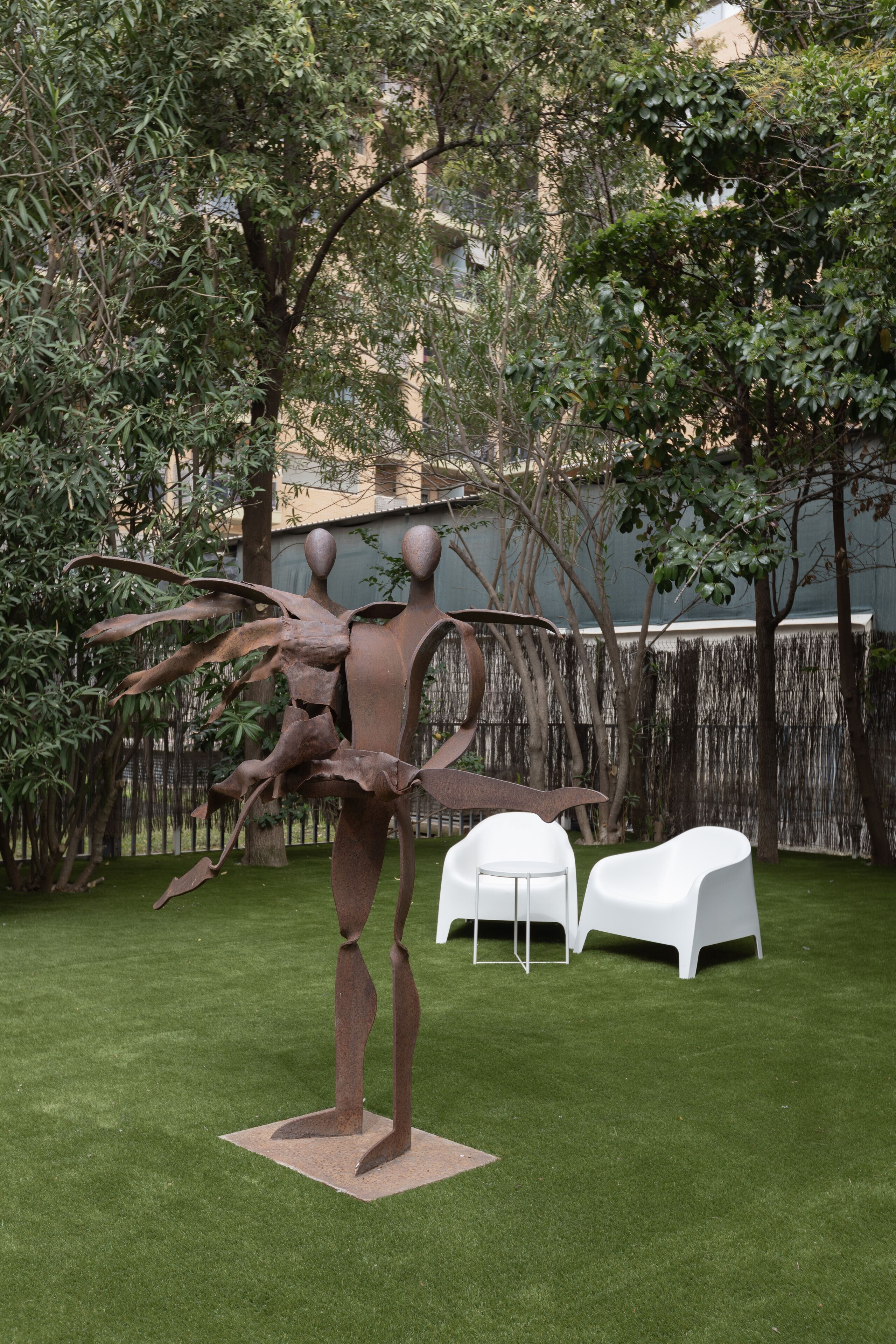 A sculpture of a human figure with outstretched arms, surrounded by a grassy area with two white chairs.