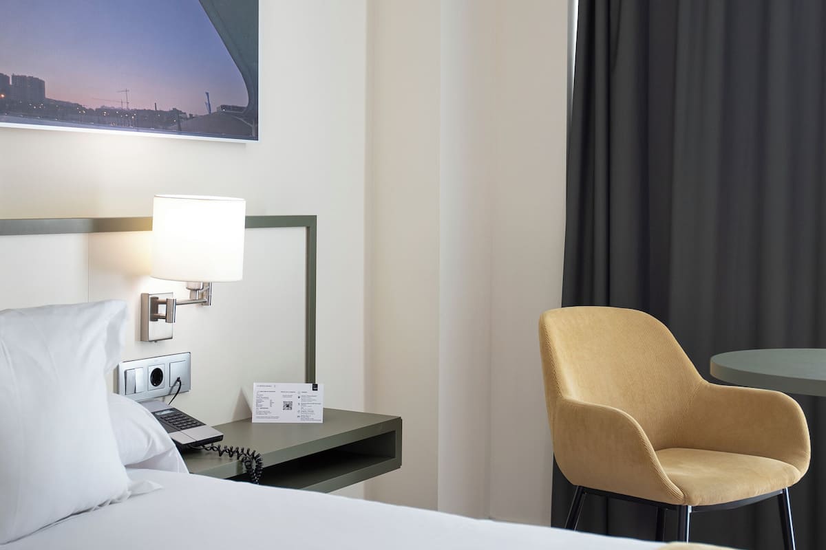 Habitación de hotel con cama, mesita de noche con lámpara, silla y vista a un paisaje urbano al atardecer.