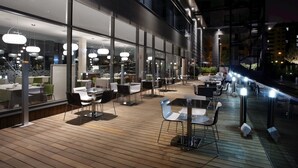 Un área de comedor al aire libre con mesas y sillas, iluminada por luces colgantes, y vistas a un edificio moderno con amplias ventanas.