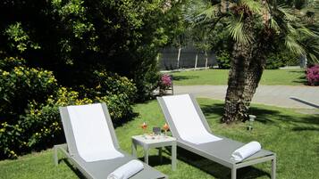 Two lounge chairs with white towels, a small table with a drink and snacks, and a palm tree in a well-maintained garden.