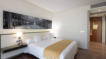 Habitació d'hotel amb un llit gran, taules de nit amb làmpades, una obra d'art penjada a la paret i una porta de fusta.
