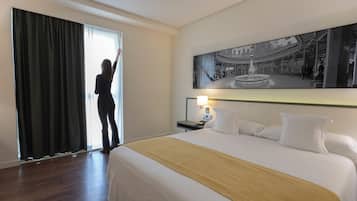 Una habitación de hotel con una cama grande, una mesita de noche con una lámpara, un cuadro en la pared y vistas a un edificio a través de la ventana.