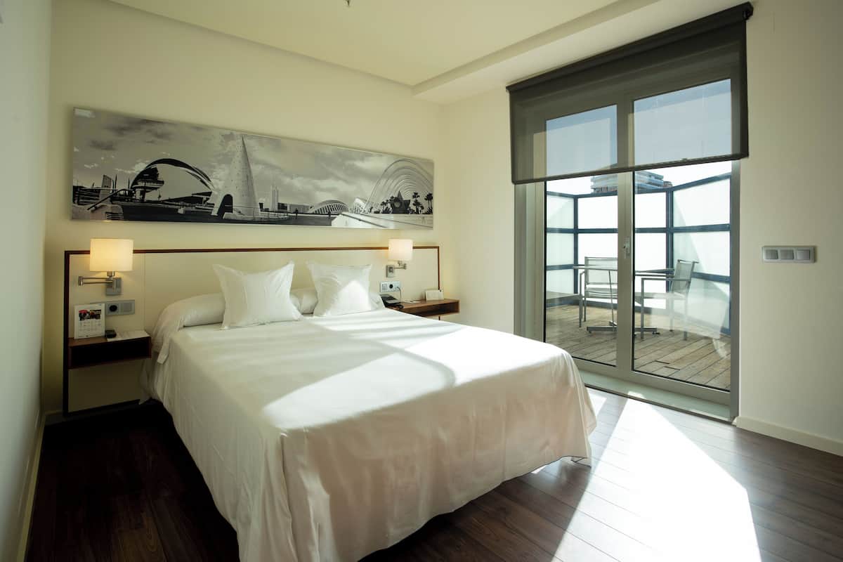 Habitación de hotel con una cama grande, un cuadro de paisaje urbano sobre la cama, un balcón con vistas y piso de madera.