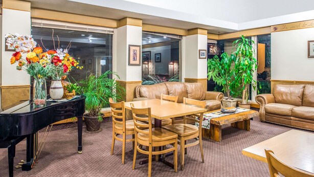 A spacious living room with a grand piano, leather sofas, a wooden dining table, and various potted plants. - Travelodge by Wyndham Kalispell (Kalispell)