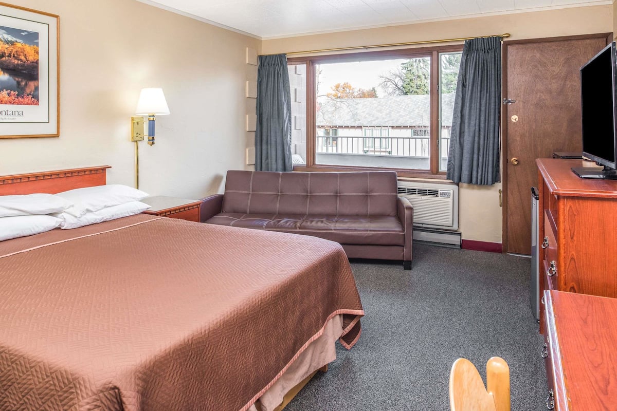 Ein Hotelzimmer mit einem Bett, einem Sofa, einem Schreibtisch mit Fernseher, einem Fenster mit Vorhängen und einem gerahmten Bild an der Wand.