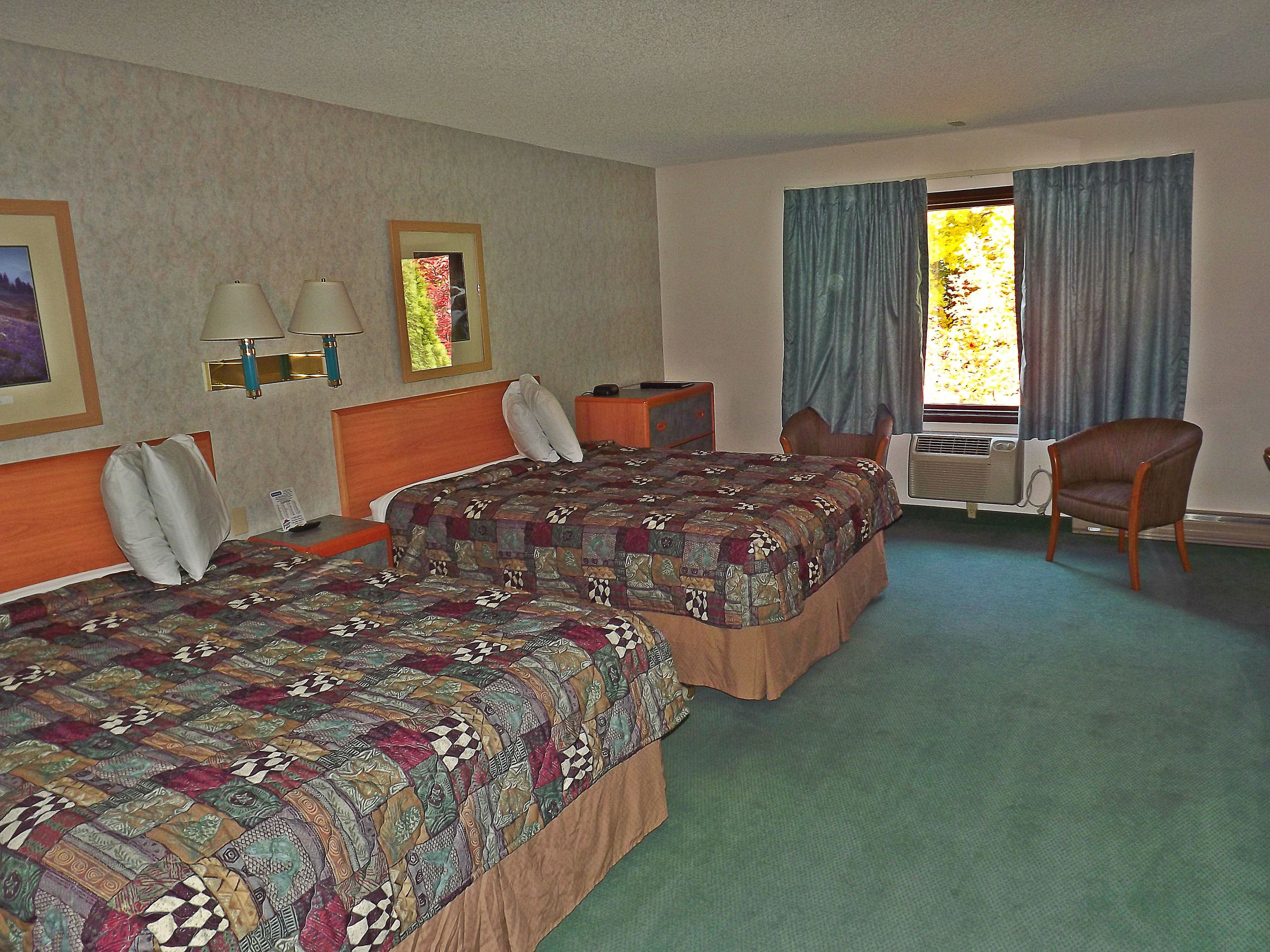 Habitación de hotel con dos camas, un escritorio, una silla, una lámpara y un ventanal con cortinas.