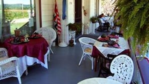 Un área de comedor al aire libre techada, con sillas y mesas de mimbre blanco, una bandera estadounidense y plantas en macetas.