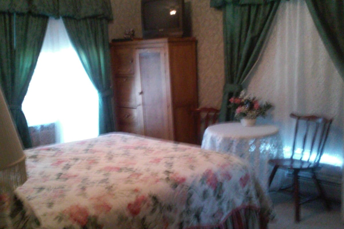 A bedroom with a bed, a dresser, a chair, a small table with a vase, and curtains.