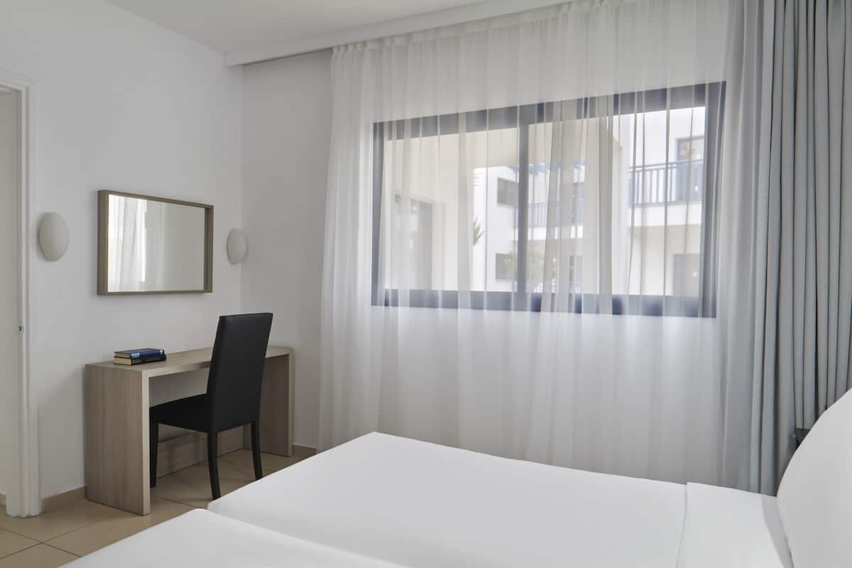 Ein Hotelzimmer mit einem Bett, einem Schreibtisch mit Stuhl, einem Fenster mit durchsichtigen Vorhängen und einem Spiegel an der Wand.
