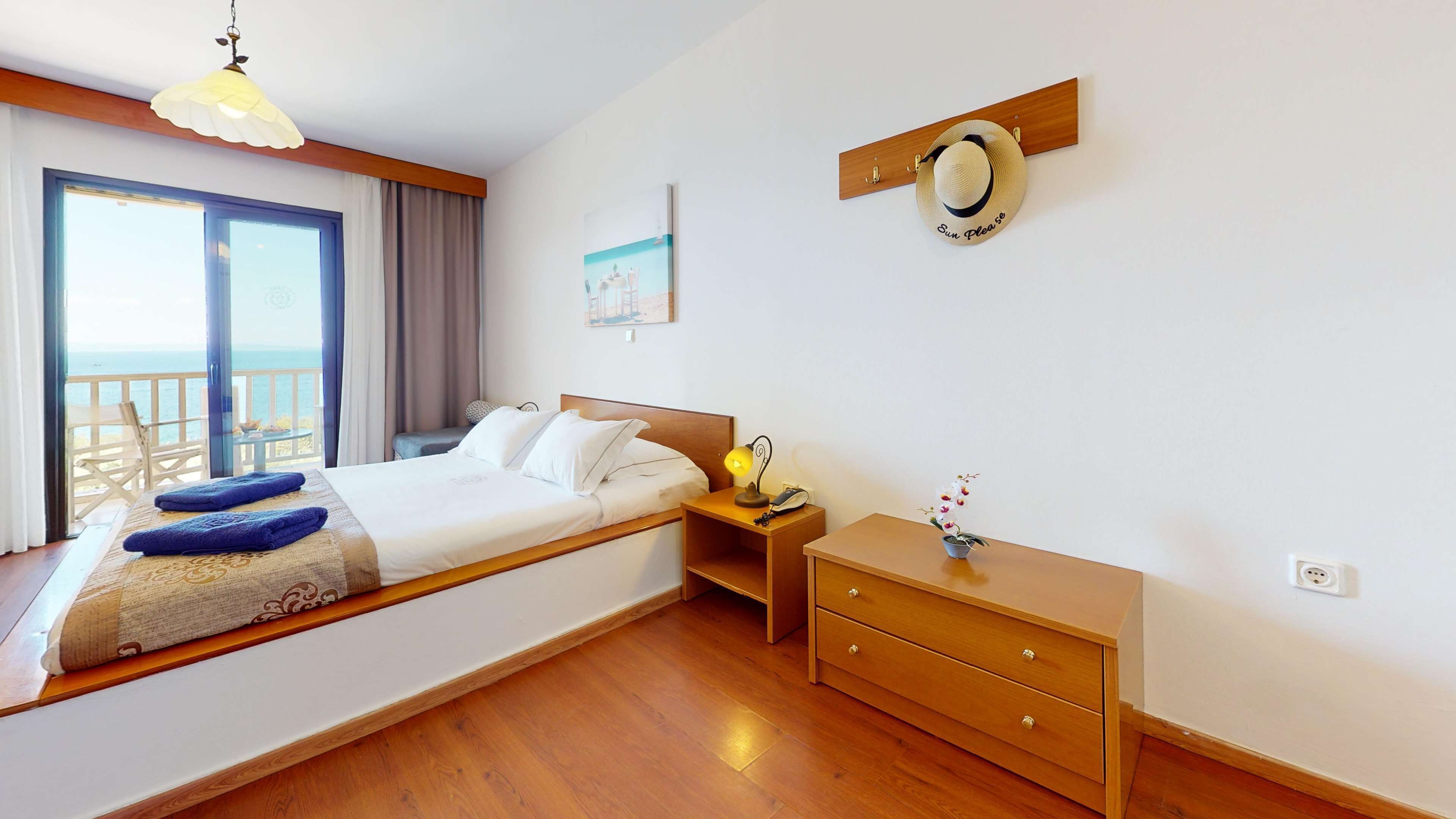 Une chambre à coucher comprenant un lit, une table de chevet, une commode et une vue sur la mer grâce à de grandes fenêtres.