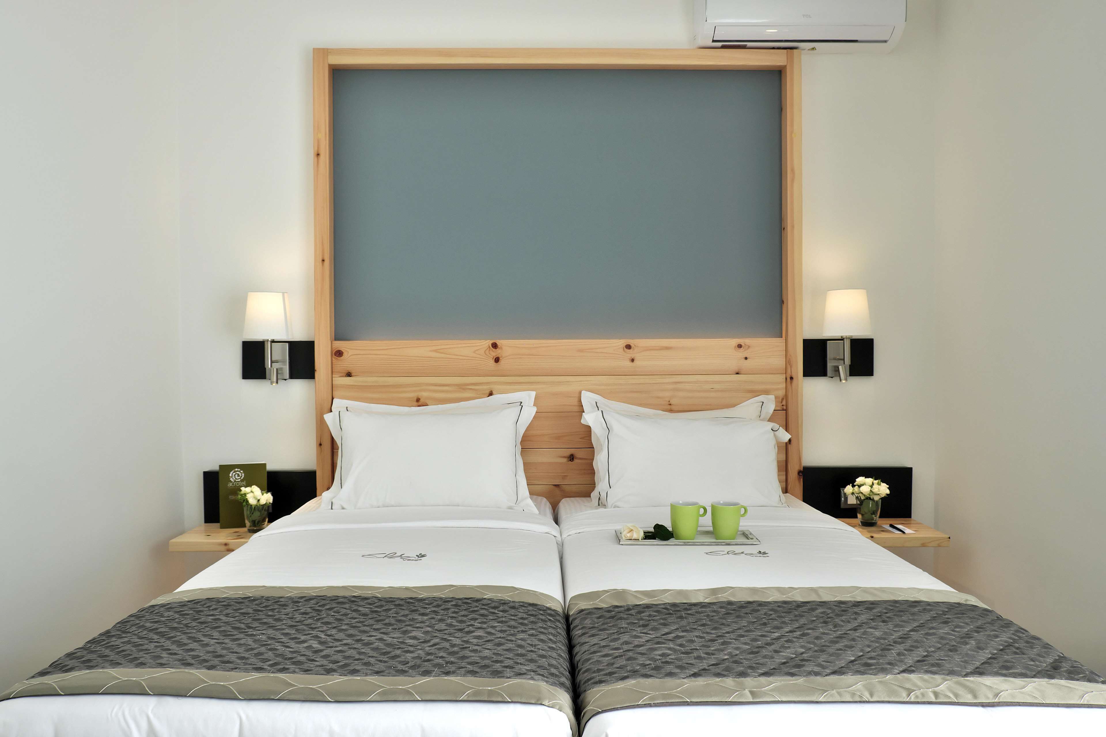 A double bed with a wooden headboard, two bedside tables with lamps, and a tray with green mugs and a white flower.