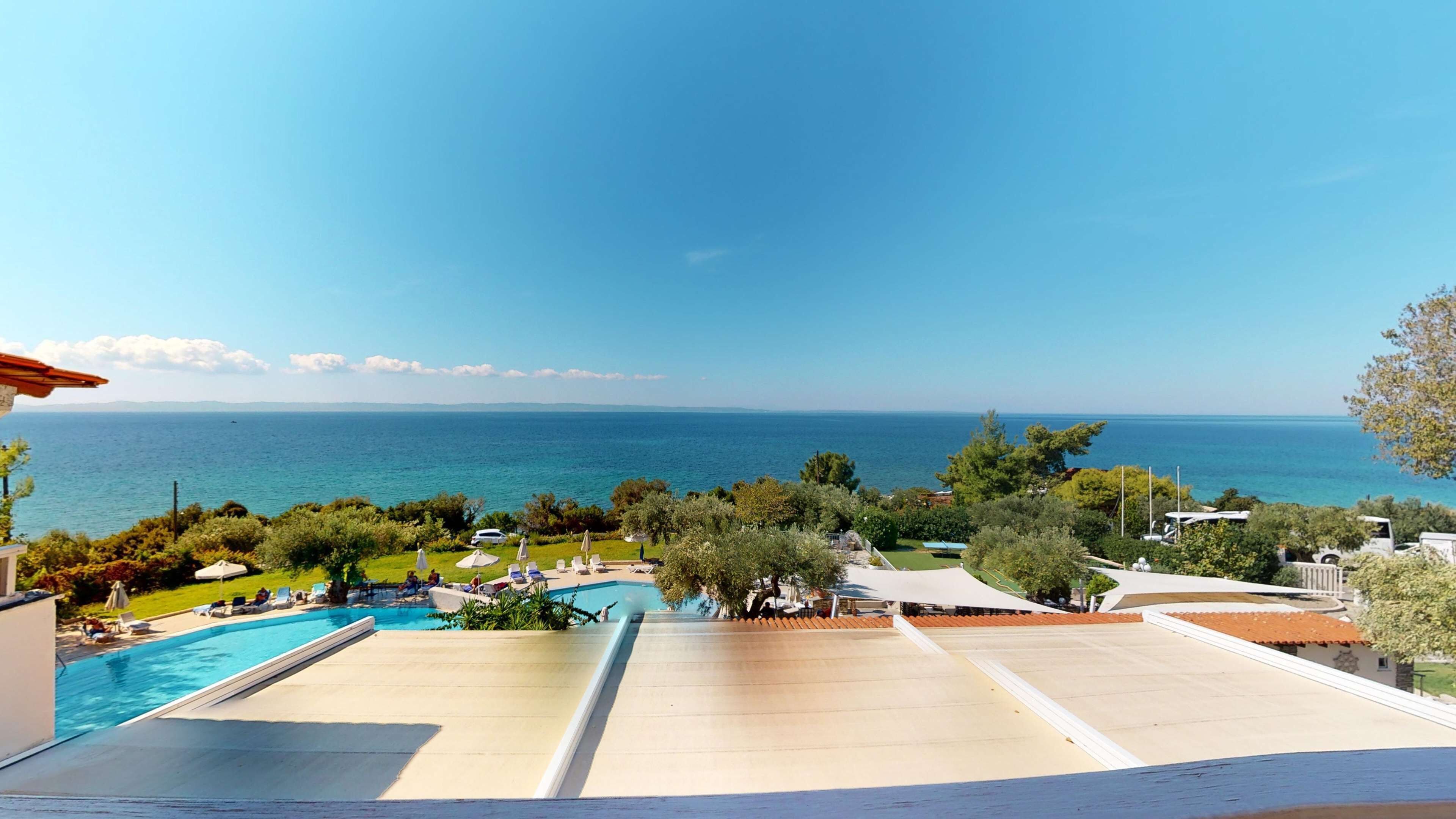 Zona de piscina con sillones de descanso y sombrillas, con vista a un mar azul cristalino y una costa distante.