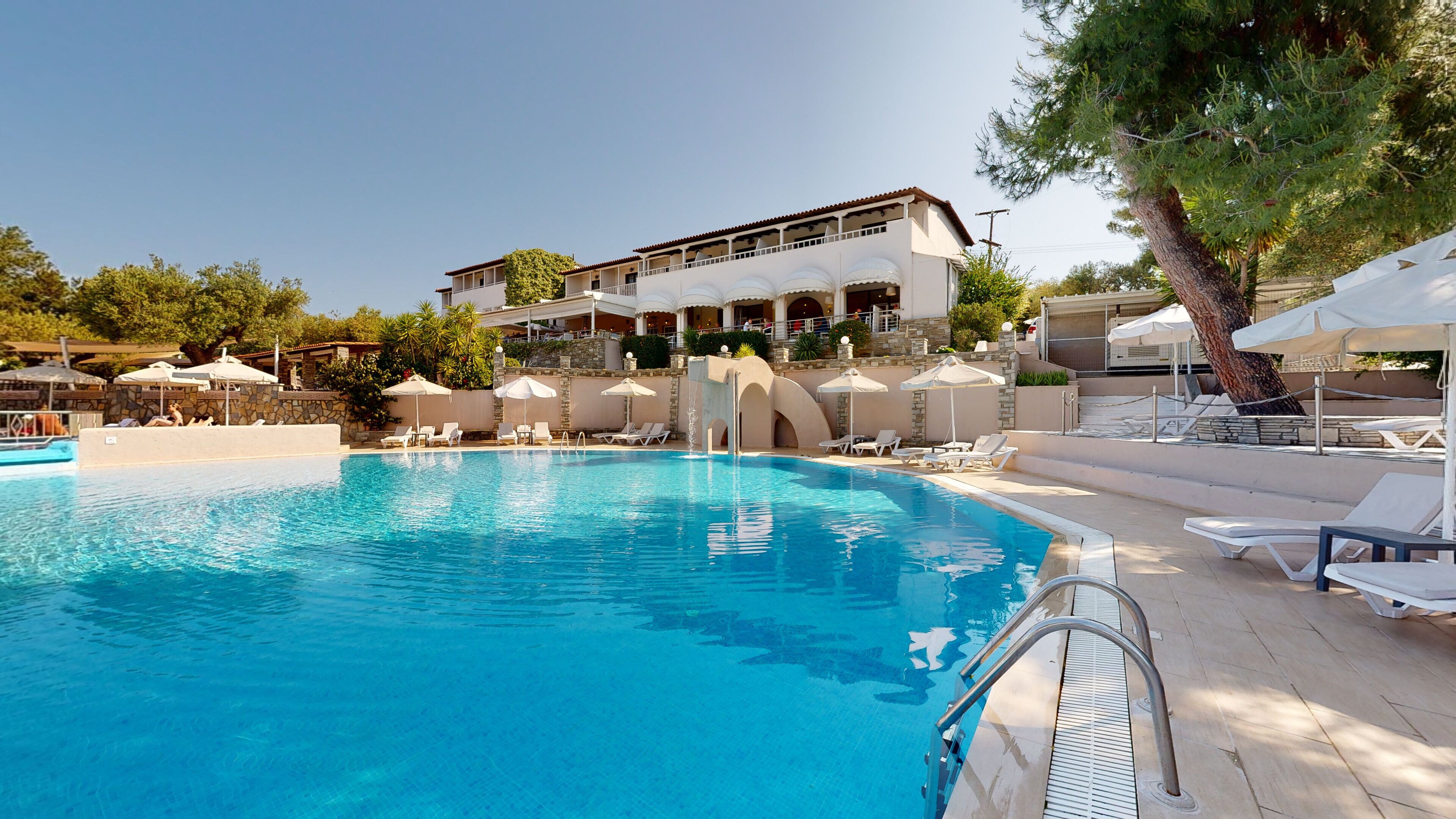 A swimming pool with lounge chairs and umbrellas, a stone wall, and a building with balconies in the background.