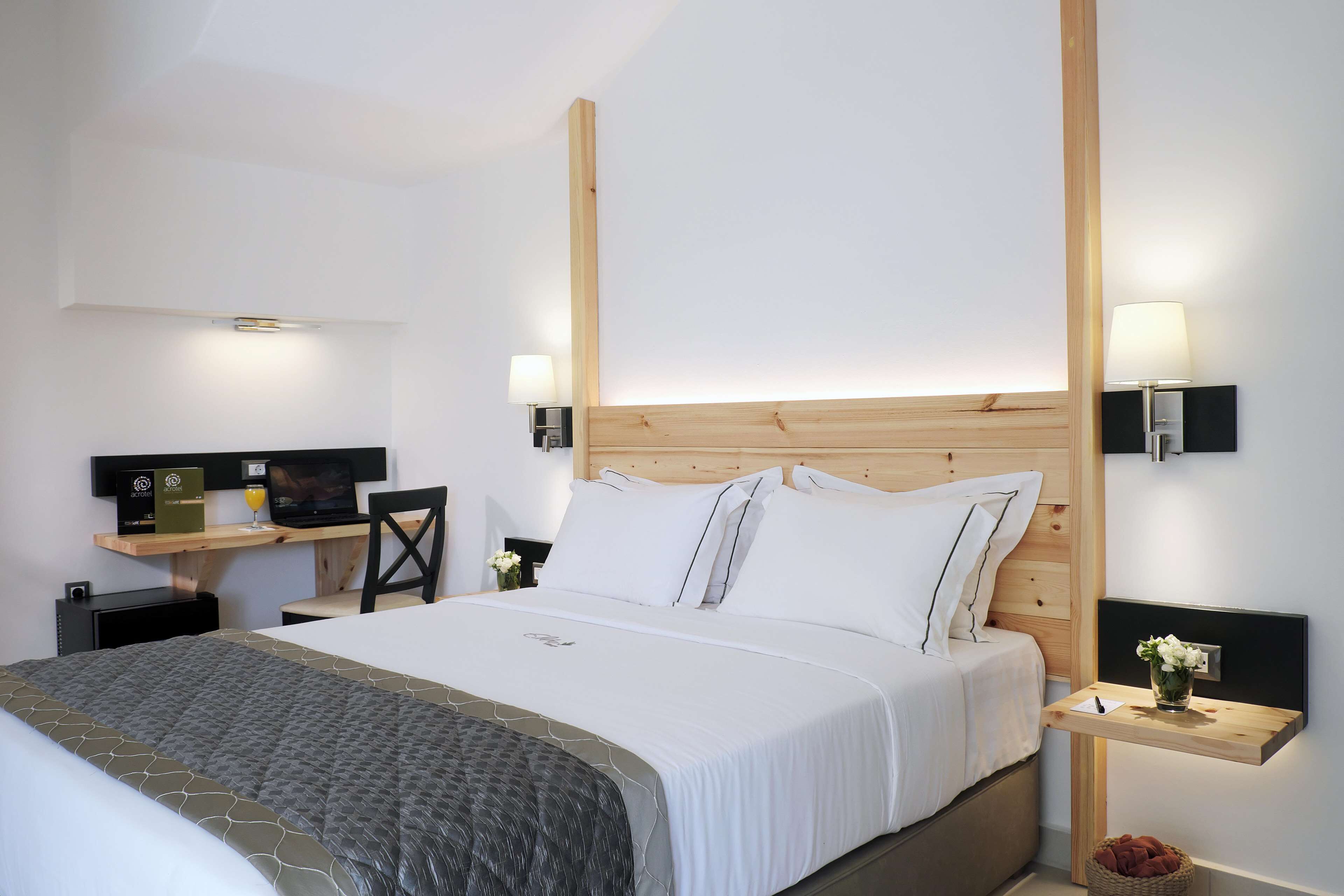 Ett modernt hotellrum med en sänggavel i trä, en stor säng med vita sängkläder, ett skrivbord med en bärbar dator och två väggmonterade lampor.