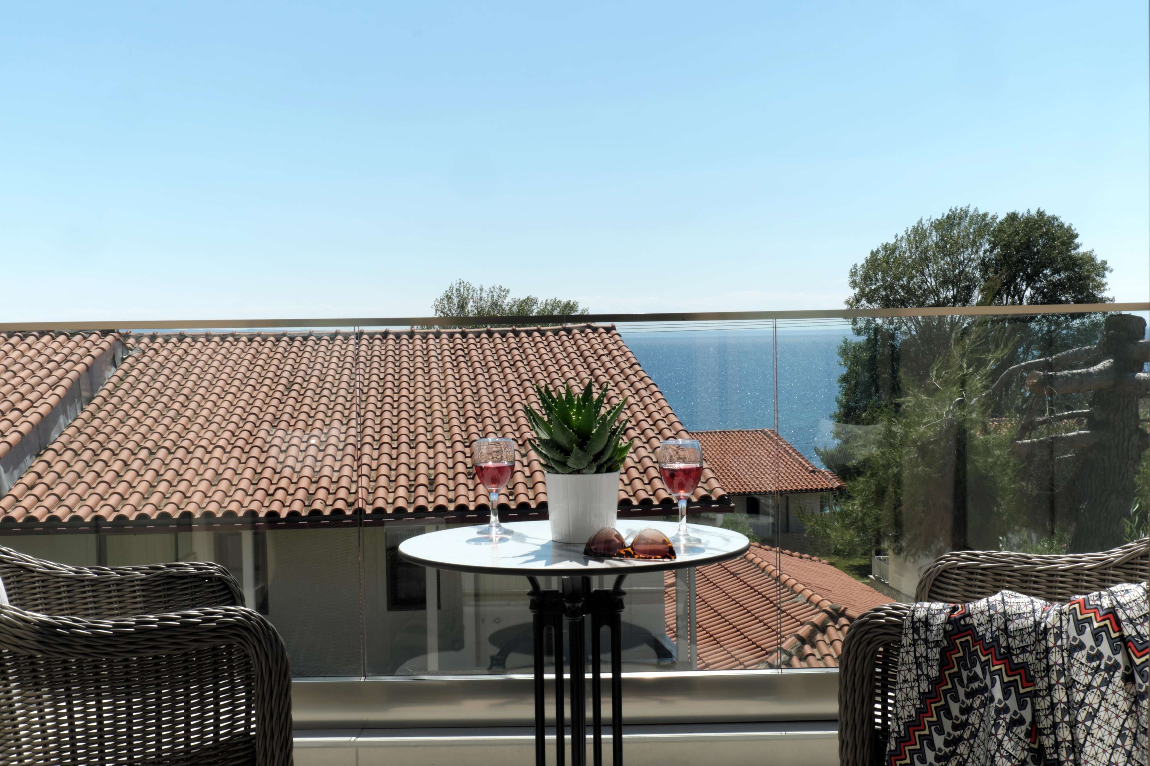 Un balcone con tetto in tegole, un tavolo con due bicchieri di vino rosso, una pianta in vaso e mobili in vimini.